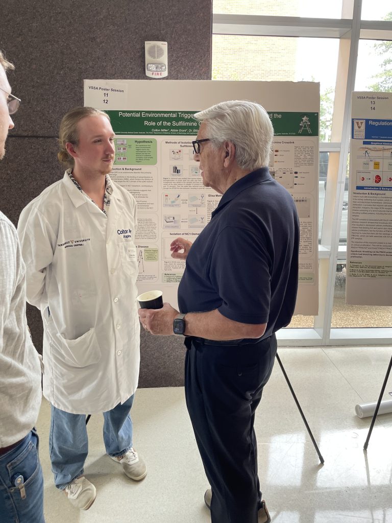 Billy Hudson talks to a student in a white coat as they both stand in front of the student's poster.