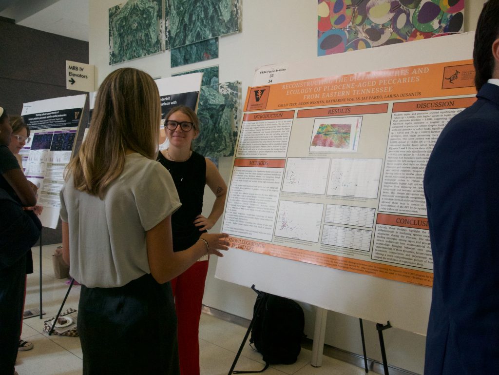 A student at a research poster talks with someone visiting their poster. The poster is branded with the Vanderbilt oak-leaf V and a kangaroo.