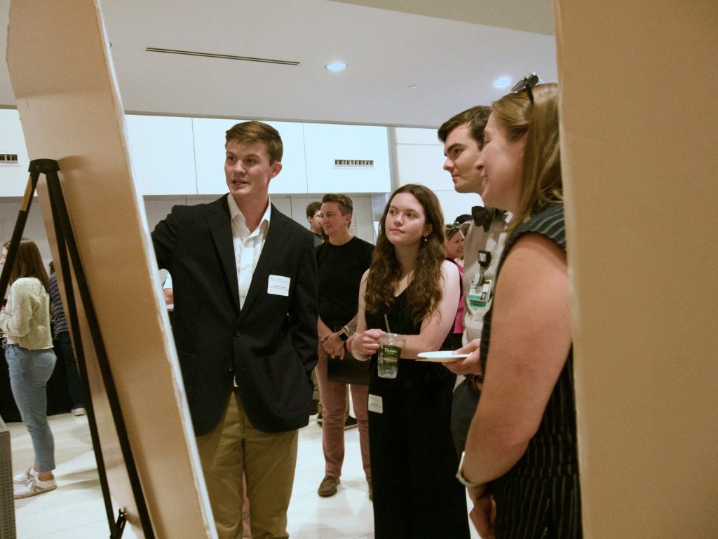 A small group of people listens to a student present on the contents of a research poster. The photo is taken partly from behind the poster.