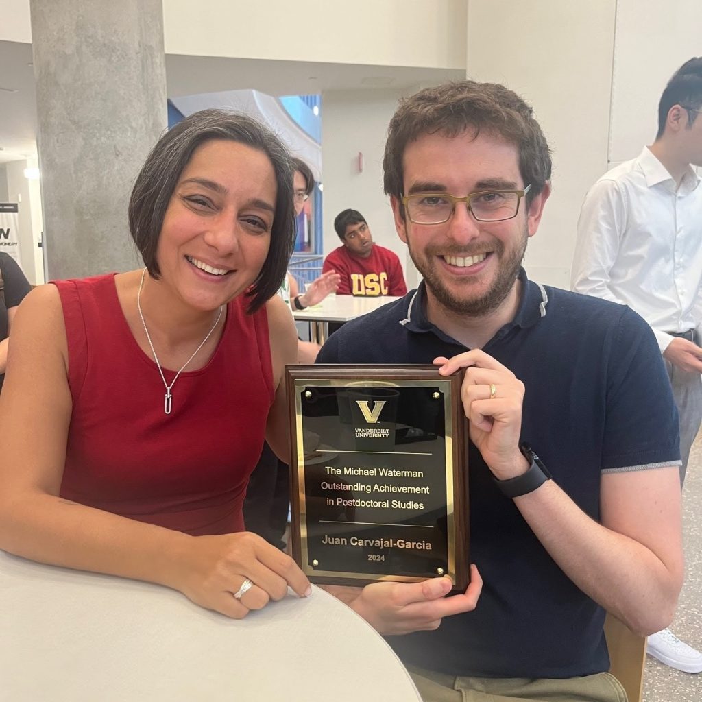 Houra Merrik (left) and Juan Carvajal-García sit at a round table and smile as Carvajal-García holds a plaque commemorating his award.