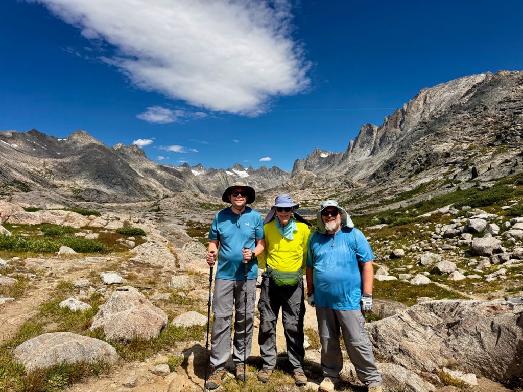 Three men in hiking gear and hats stand in front of a rocky landscape. The sky is mostly clear and bright blue except for a stray, fluffy cloud on the left side. From left to right, the men are Mike Goodman, Chuck Sanders, and Wade Van Horn.