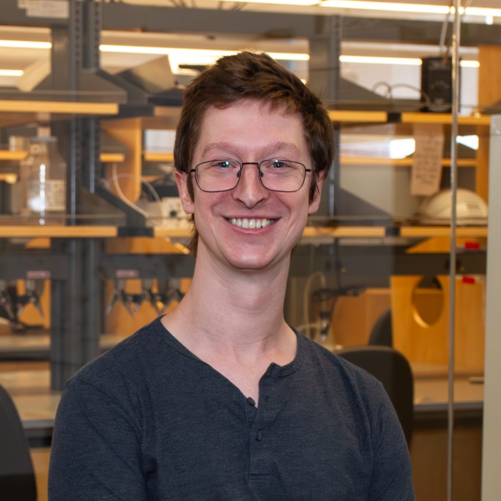 Headshot of Benjamin Brown. He's in front of a glass window. You can see a lab bench and shelves behind him.