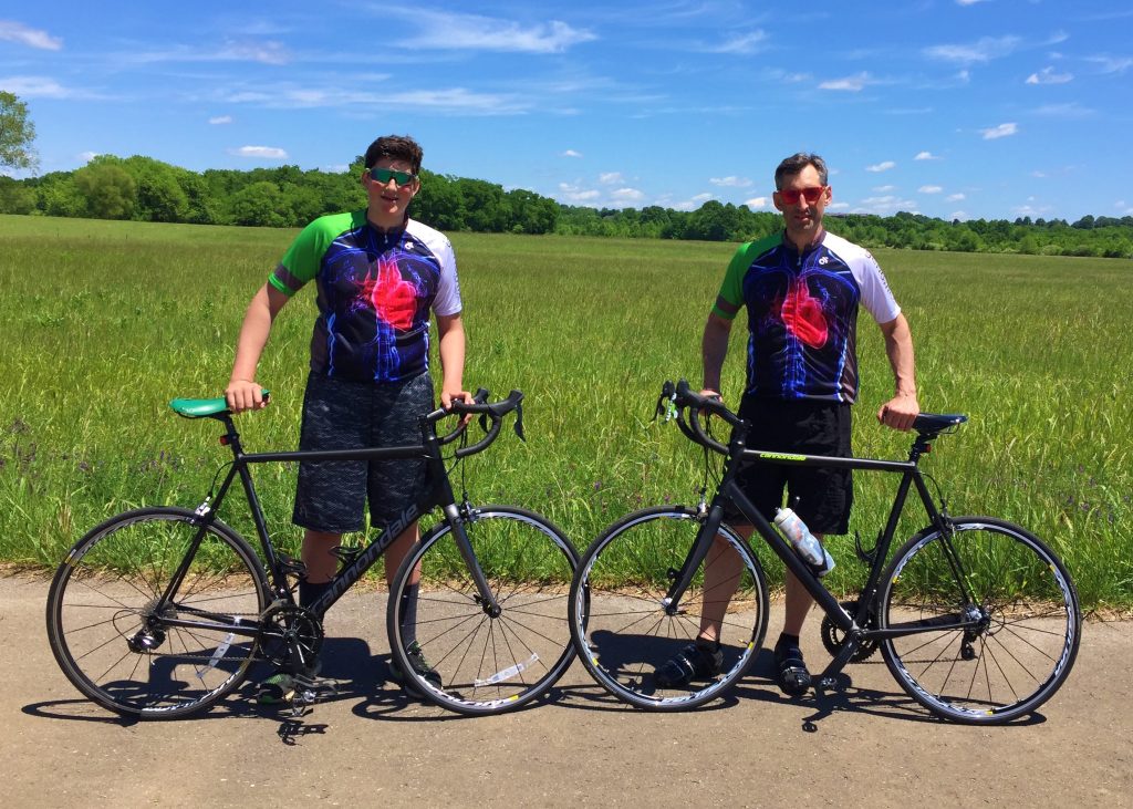 David Vigerust (right) and his son posing behind their bicycles in front of a sunny, grassy field. They're in matching cycling shirts.