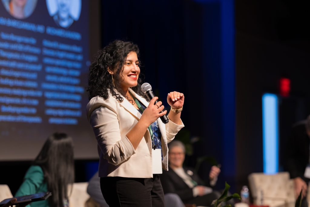 Kavya Sharman, in a white blazer, holding a microphone and smiling at her audience. In the background, out of focus, you can see a projector screen and three people seated in front of it, as if they were a panel of speakers.