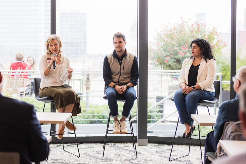 Three people sitting on stools in front of a wall of windows. The person on the left is holding a microphone. Kavya Sharman, in jeans and a white blazer, is seated on the right-most chair. City buildings, greenery, and an outdoor seating area can be seen through the window.