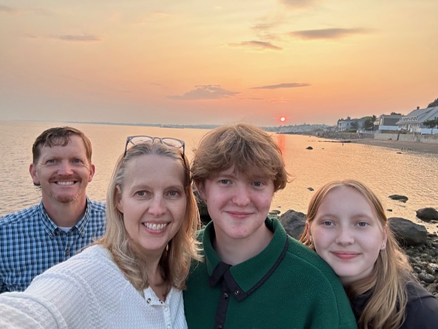 Selfie that Lori took of her family. From left to right are J.J., Lori, Cole, and Olivia. They are on a rocky beach or outcropping in front of a bay at sunset.