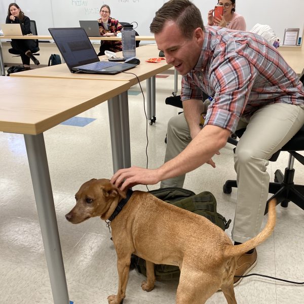 A man pets a dog in a classroom while others sit at a large table