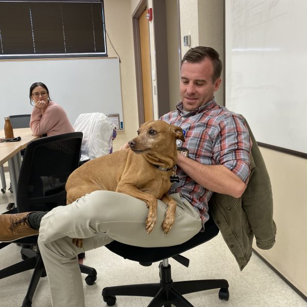 A dog sits on a man's lap as a classmate looks on
