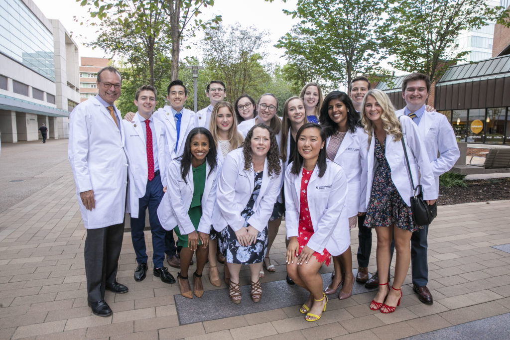 A large group of smiling VUSM students and faculty pose for a photo outdoors wearing their white coats.