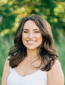 A woman in a white top stands outside and smiles