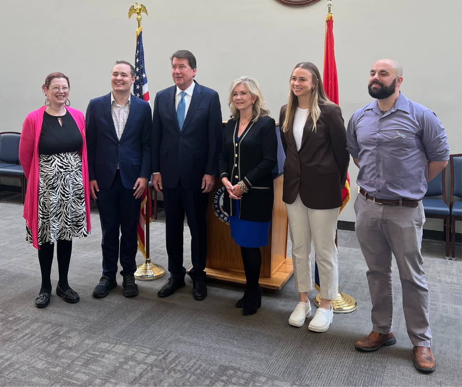 A group of men and women gather in front of a lectern and flags to pose for a photo.