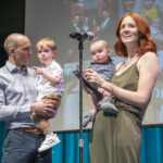 A man and a woman both hold baby boys while standing on a stage.