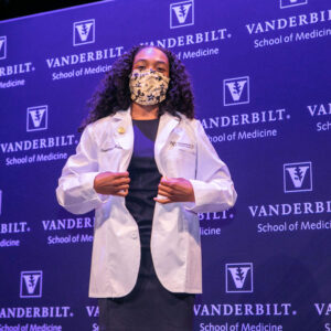 A woman in a mask and white coat stands in front of a medical school backdrop