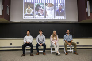 Three men and one woman sit in chairs at the front of a room below a screen with their photos