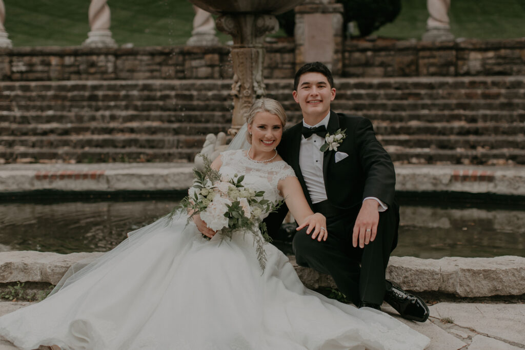 A newlywed couple takes a photo in their wedding clothes in front of a fountain