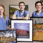 Three medical students in scrubs pose with framed artwork