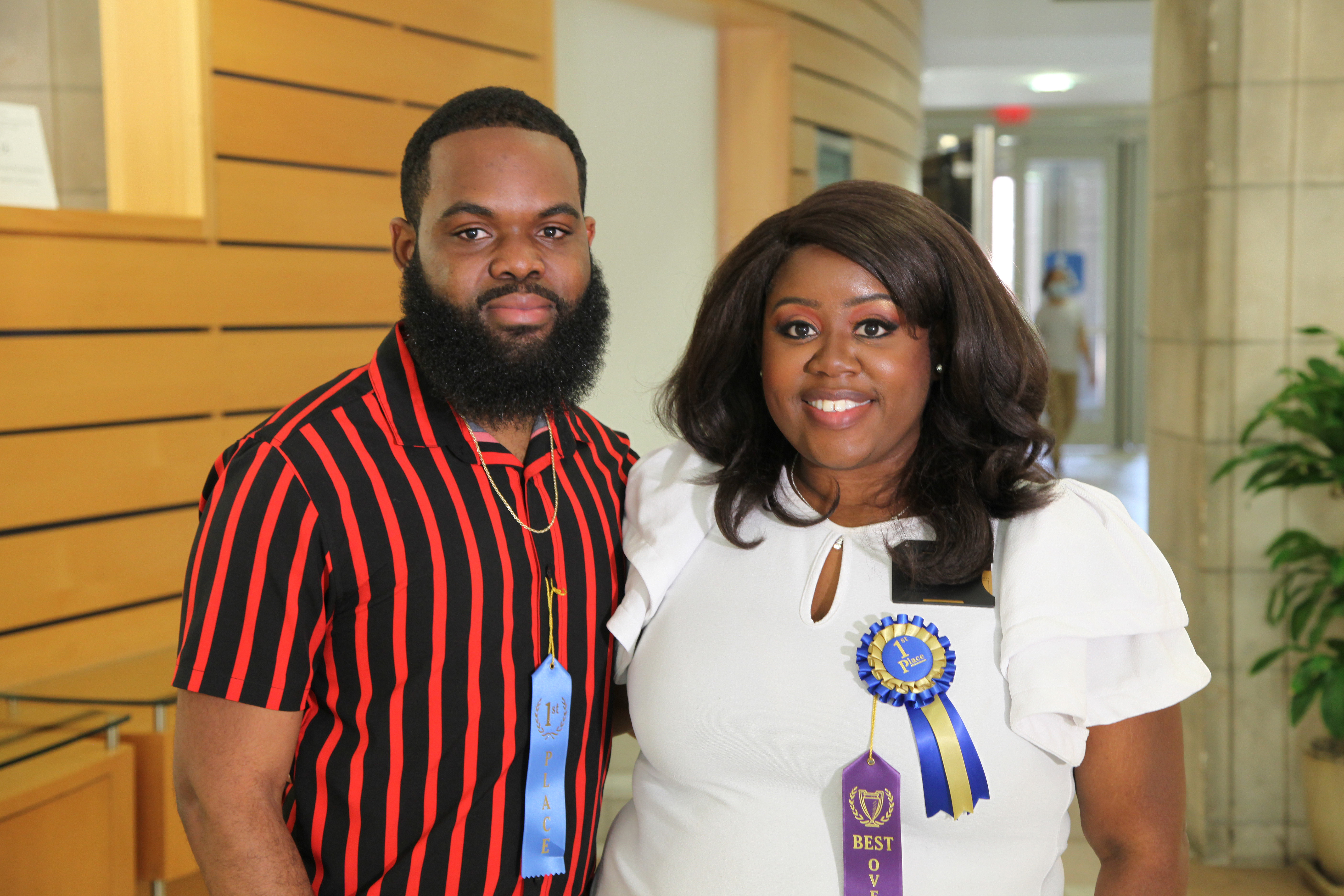 Carlan Romney and Jessica Thomas, doing a side hug for the photo and wearing ribbons that say “1st Place” (both) and “Best overall” (Thomas). Romney is wearing a black and red striped shirt and Thomas a white blouse. They’re standing in the atrium of MRBIII on the Vanderbilt campus.