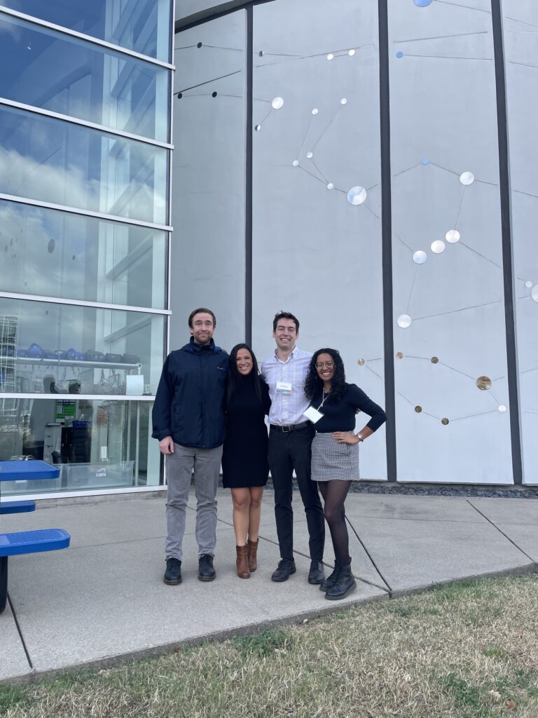 Two men and two women pose for a photo outdoors in front of a window and a mosaic.