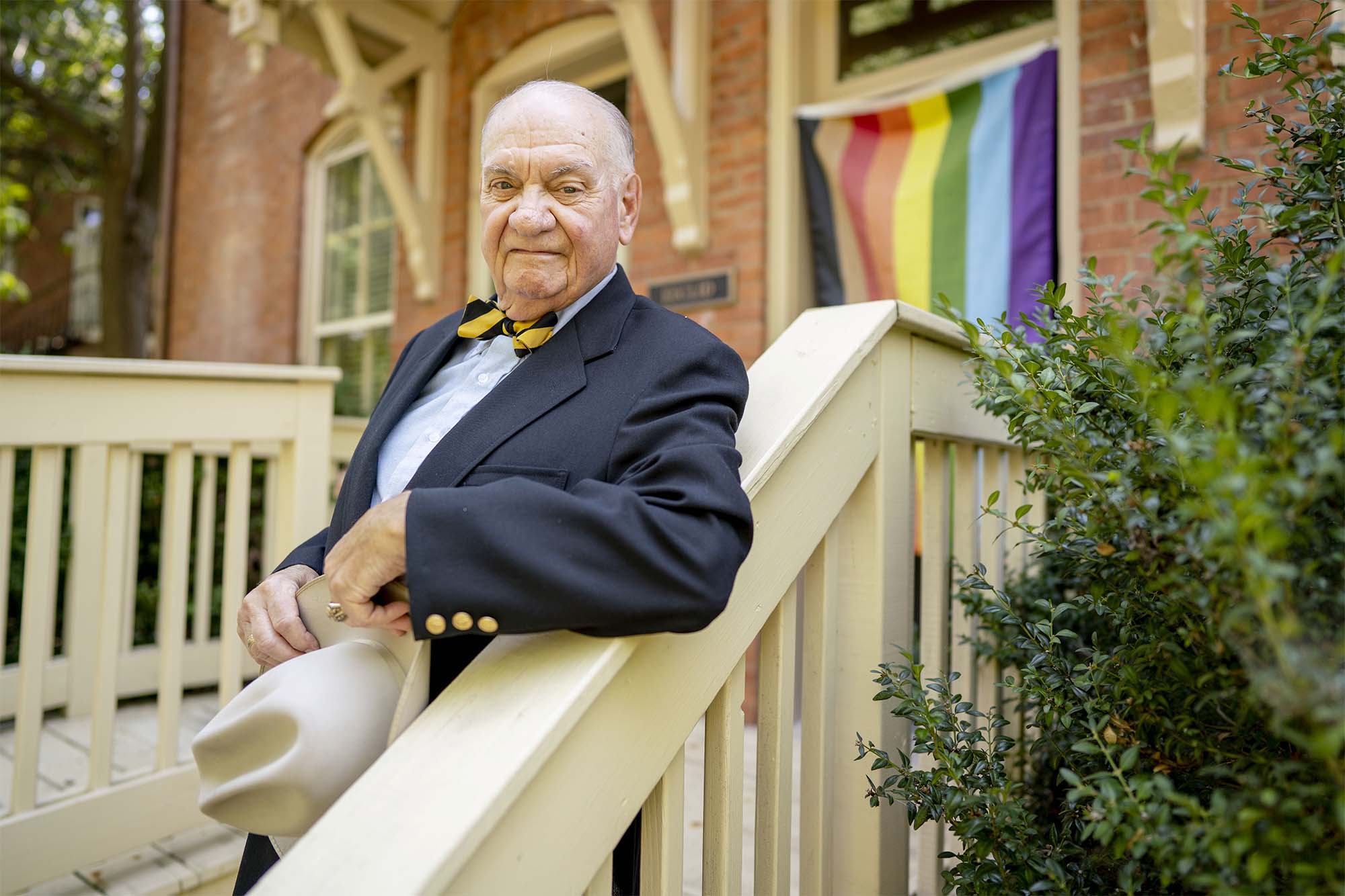 K.C. Potter, an older white man in a suit with a bowtie and holding a hat, leans on the stair railing outside the Center. A pride flag is visible in the background.