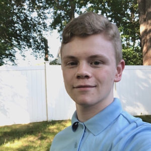 Undergraduate writing consultant Cameron Sheehy pictured wearing a light blue polo shirt, white fence and tree foliage in the background