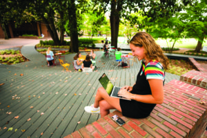 Female student sitting on the brick wall outside of Rand Hall working on a laptop with a group of people sitting in the background