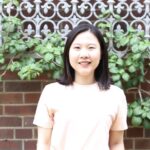 Asian woman with shoulder length hair standing in front of brick wall with white patterned fence and ivy