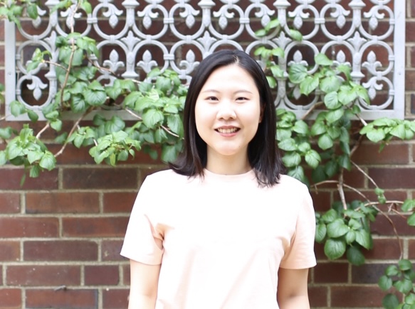 Asian woman with shoulder length hair standing in front of brick wall with white patterned fence and ivy  