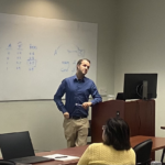 Man in blue shirt and khaki pants stands at lectern in front of whiteboard with statistical lesson
