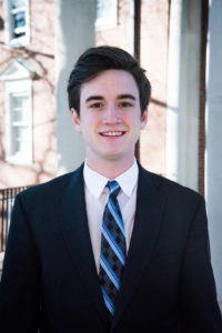 A color photograph of Ryan Connor, a young white man with neat dark hair standing in front of a sunlit brick building. He is wearing a dark suit, a white button down shirt, and a blue striped tie.