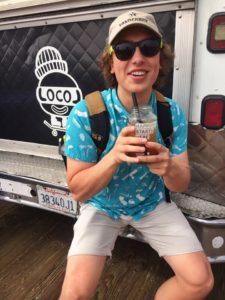 A color photograph of Tarin Denney, a young white man with shaggy brown hair. He is sitting on the back bumper of a white vehicle with a black logo saying "Locol" and holding a plastic drink with both hands. He is wearing a bright blue patterned shirt, pale khaki shorts, black sunglasses, a tan baseball hat, and a tan and black backpack.