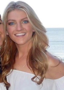 A color photograph of Paige Oxley, a smiling young white woman with long loosely curled blonde hair. She is wearing an off-the-shoulder white top and is standing outside in front of water. Another person is cut off of the left side of the image.