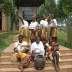 Photograph of seven African women wearing traditional dress and posing outside a brick building while holding instruments. Picture taken in Uganda.