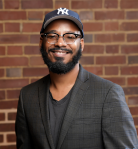 Headshot of Webster Heath. Subject is wearing a New York Yankees cap and professional dress. 