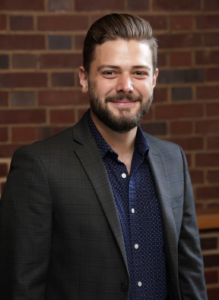 Headshot of E. Kyle Romero. Subject is wearing a blue shirt and black blazer.