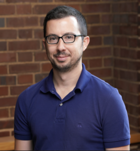 Headshot of Daniel Genkins. Subject is wearing a blue shirt and standing in front of a brick wall.