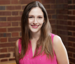 Headshot of Katherine Mckenna. Subject is wearing a pink shirt and is standing before a brick wall.