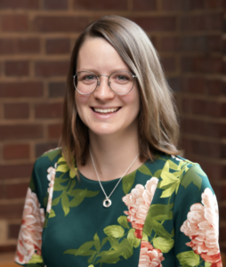 Headshot of Elizabeth Barma. Subject is wearing a green dress and standing in front of a brick wall.