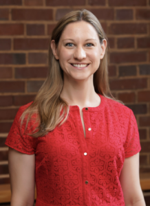 Headshot of Melanie Forehand. Subject is wearing a red dress and is standing in front of a brick wall.