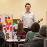 Kevin Galloway stands next to a board covered in yellow, pink and orange sticky notes. He has a presentation remote in one hand as he lectures to students seated at tables and chairs.