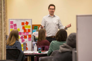 Kevin Galloway stands next to a board covered in yellow, pink and orange sticky notes. He has a presentation remote in one hand as he lectures to students seated at tables and chairs.