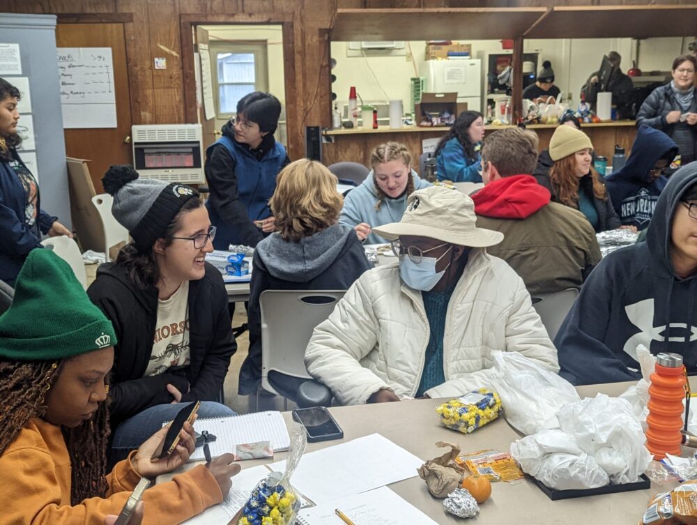 A group of people gathered around a table with papers and bags of popcorn. In the background, a second table with more people gathered around talking about evolution.