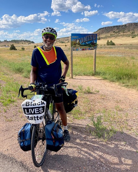Scott Edwards posing in front of the Welcome to Wyoming sign. Wearing a bike helmet and a flourescent orange bib while standing over his bike. Green grass and hills in the background with blue sky and white clouds