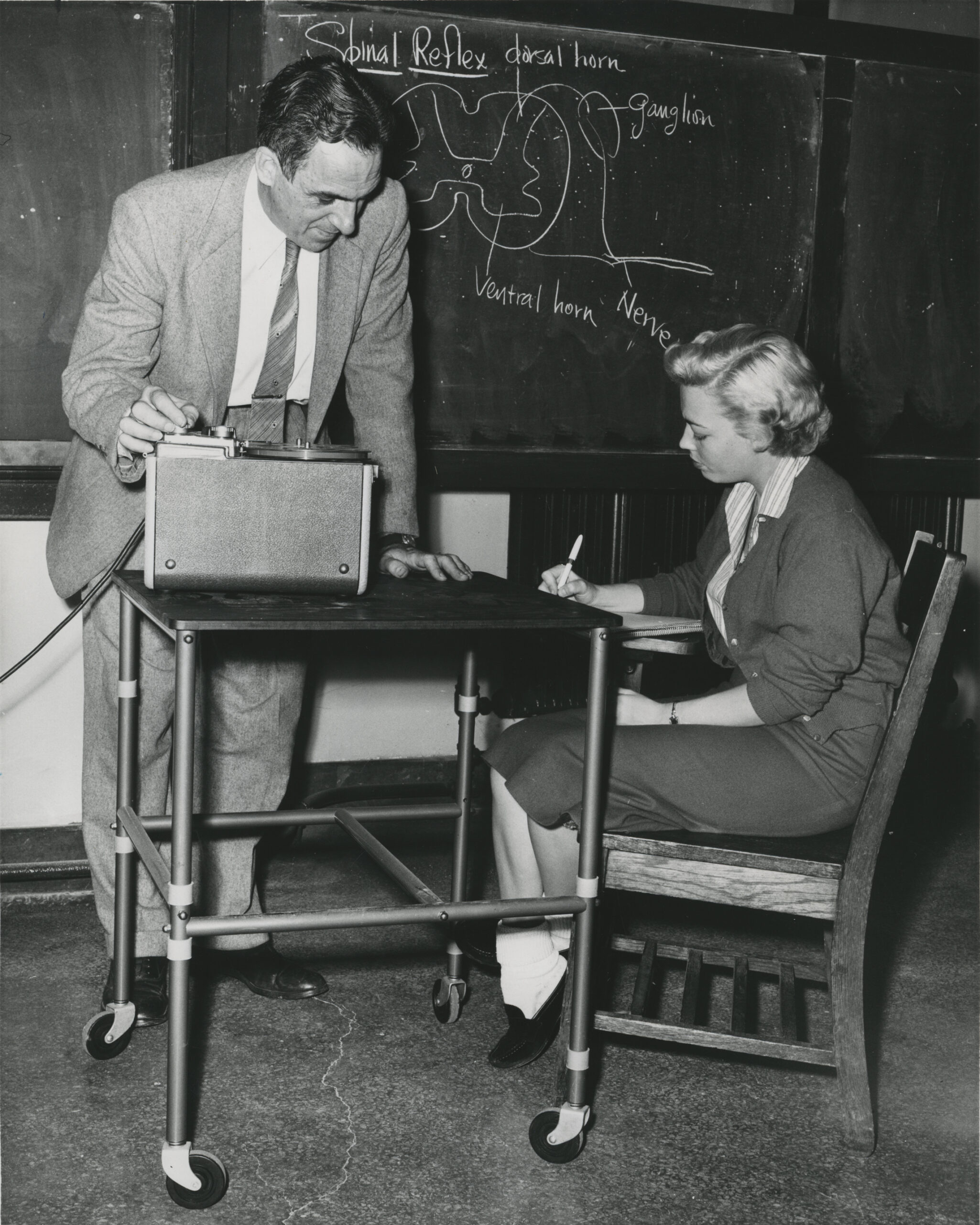 Claude in a suit and tie with a chalkboard behind him helping a student