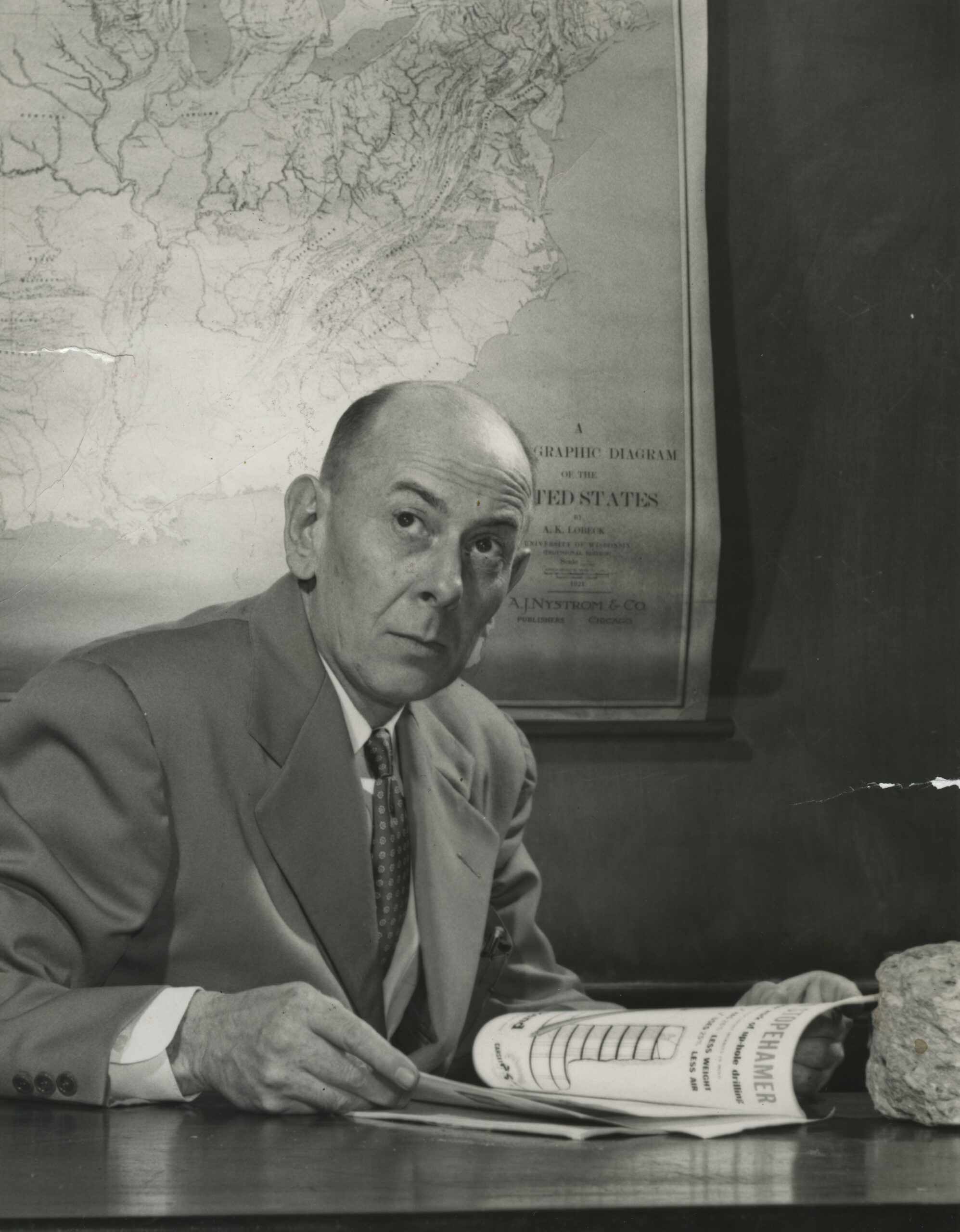 Willard Jewell looking up from his desk with a map behind him, greyscale.