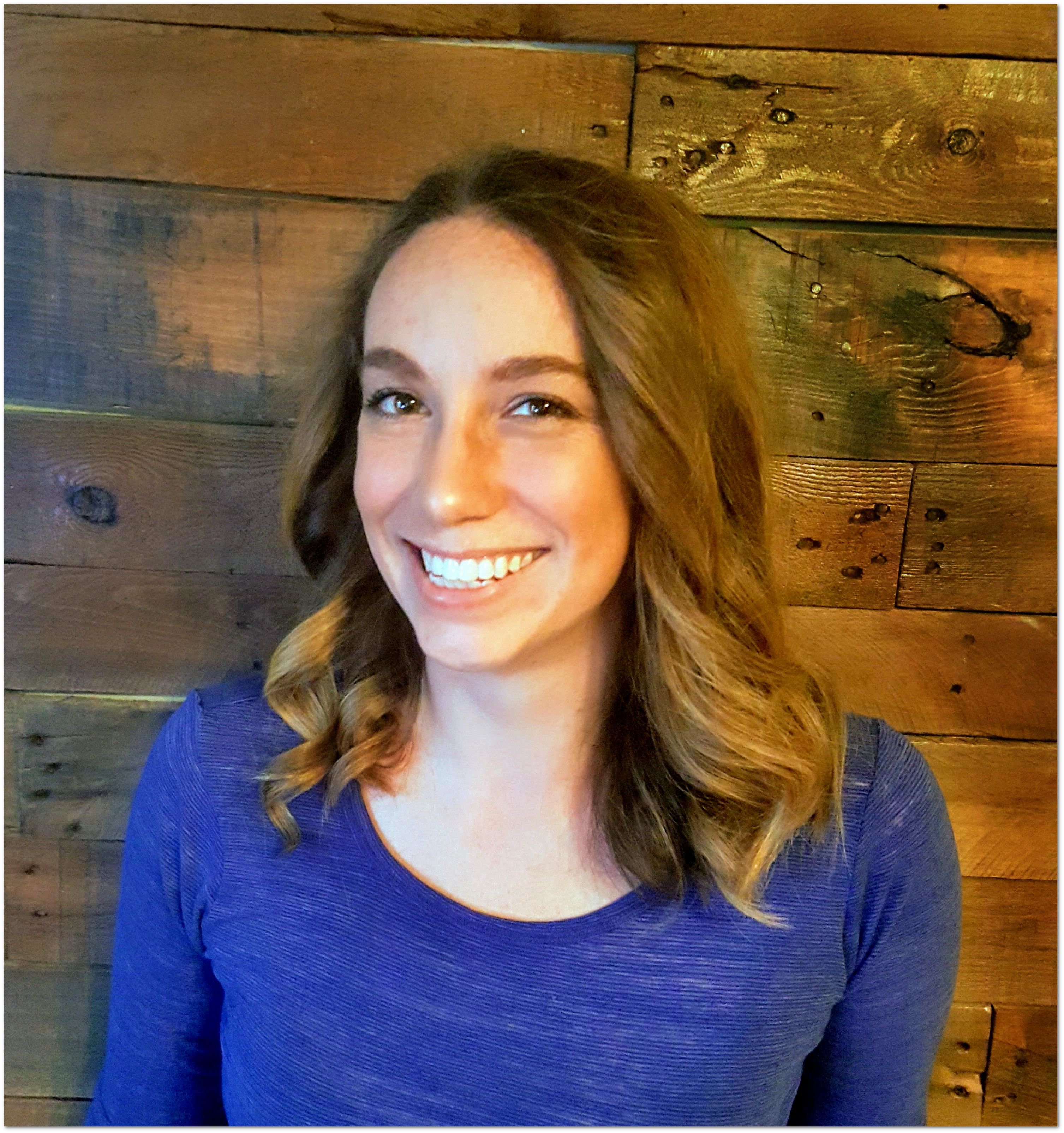 Abbe LaBella smiling big for her headshot while wearing a blue shirt. She's standing in front of a wood plank wall.