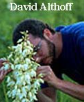 A man with short dark hair and a beard, closely examining a cluster of white flowers. He is wearing a blue shirt and appears to be outdoors in a grassy area. The caption reads 