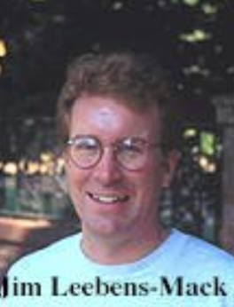 A man with short, light brown hair and glasses, smiling. He is wearing a light-colored t-shirt and is outdoors with blurred foliage in the background. The caption reads 