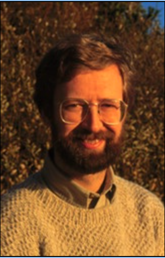 A man with glasses and a beard, wearing a sweater, smiling. He is outdoors with a background of trees and foliage.