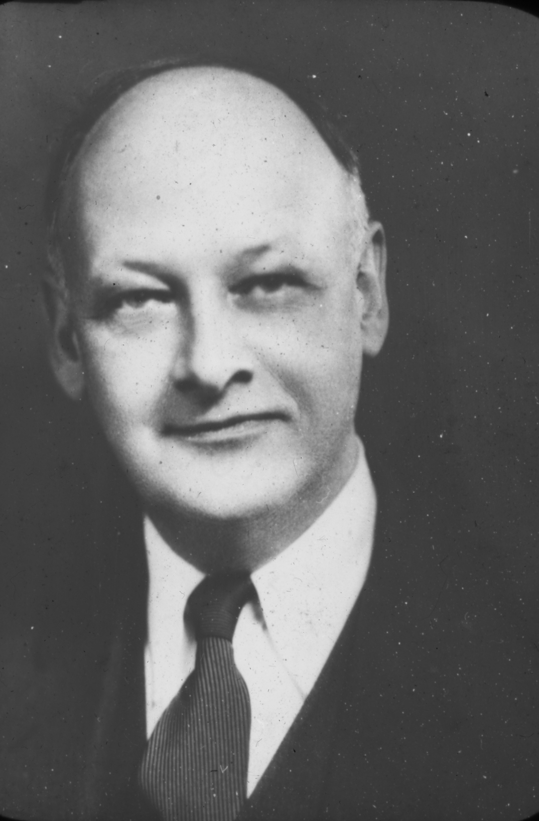 A black-and-white portrait of a middle-aged man with a bald head, wearing a suit and tie. He has a slight smile and is looking directly at the camera. The background is plain and dark, highlighting his face.