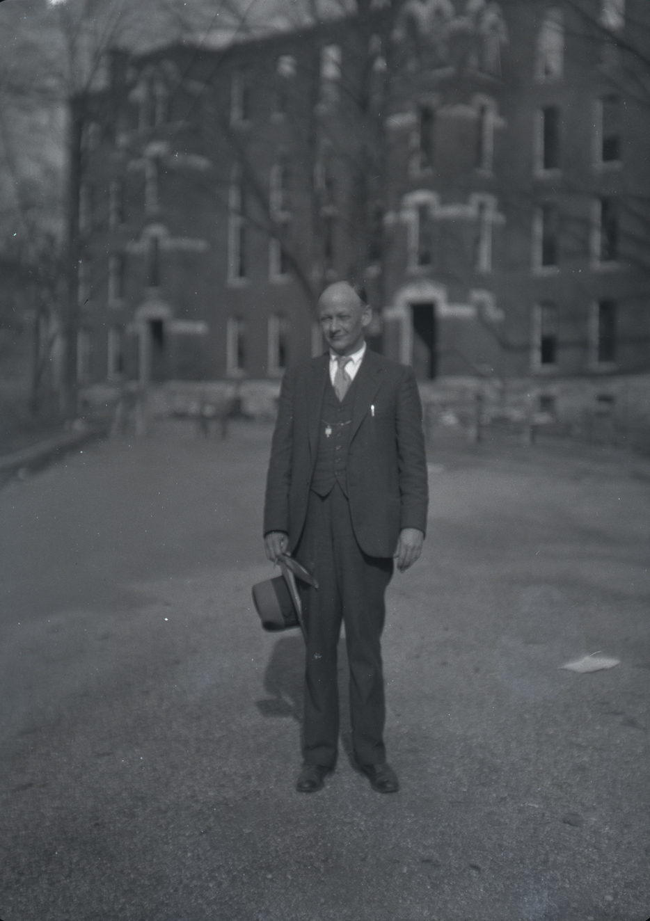 A black-and-white photograph of Reinke standing outdoors in front of a large, multi-story brick building. He is wearing a suit, tie, and vest, holding a hat in his left hand. The background is slightly blurred, focusing on the man.